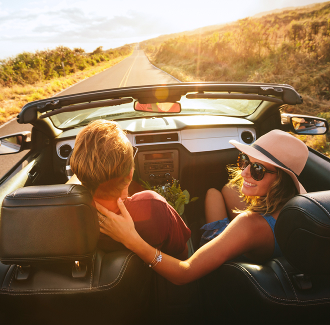 Smiling couple driving in a convertible on an open road at sunset, enjoying a scenic road trip.