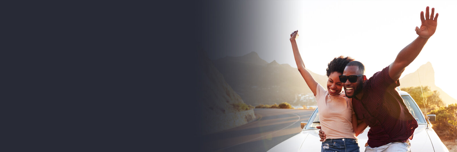 Smiling couple standing in front of a car on a scenic road, raising their arms joyfully at sunset